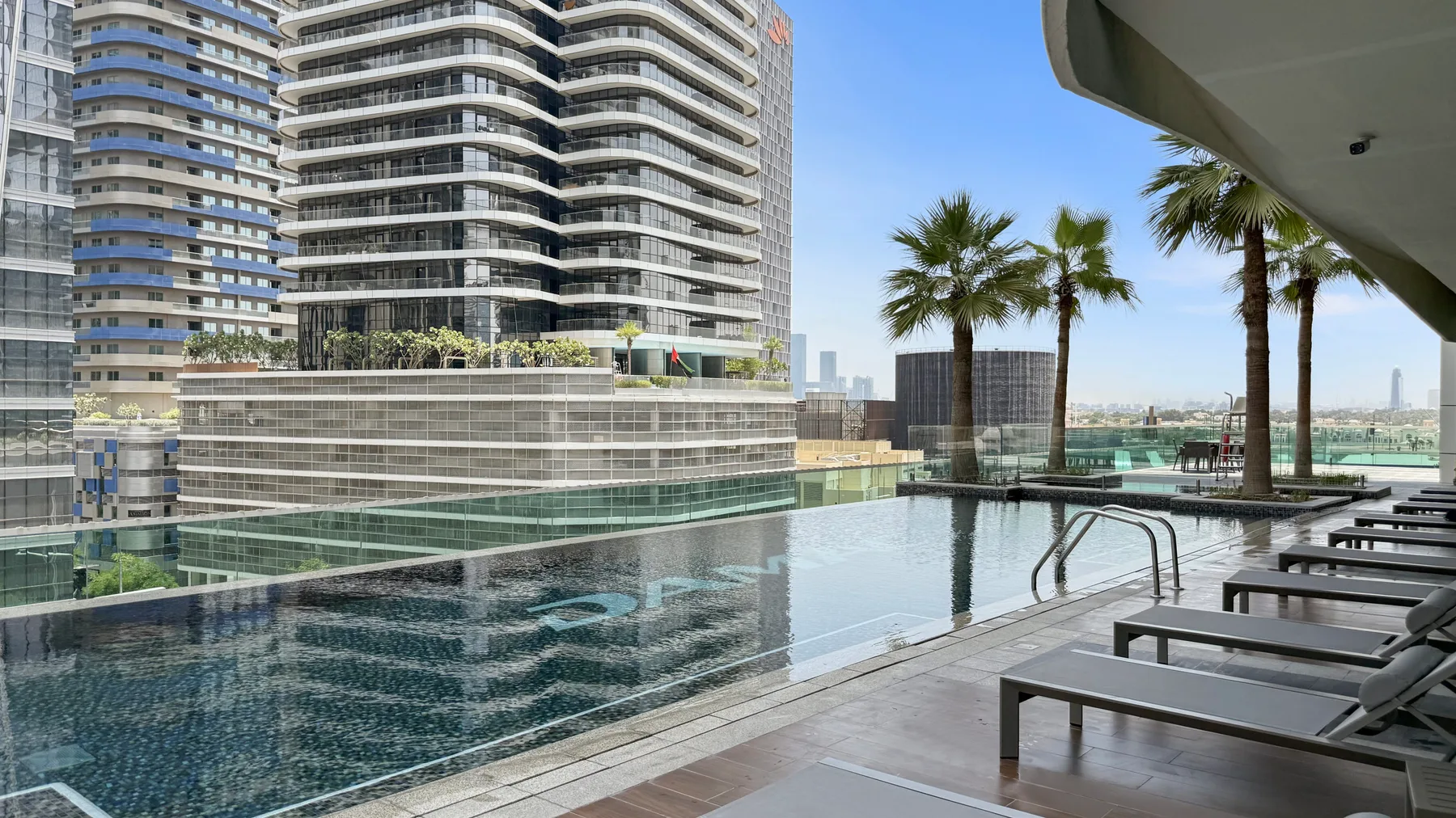 Una piscina en la azotea junto a edificios modernos, con palmeras y tumbonas.
