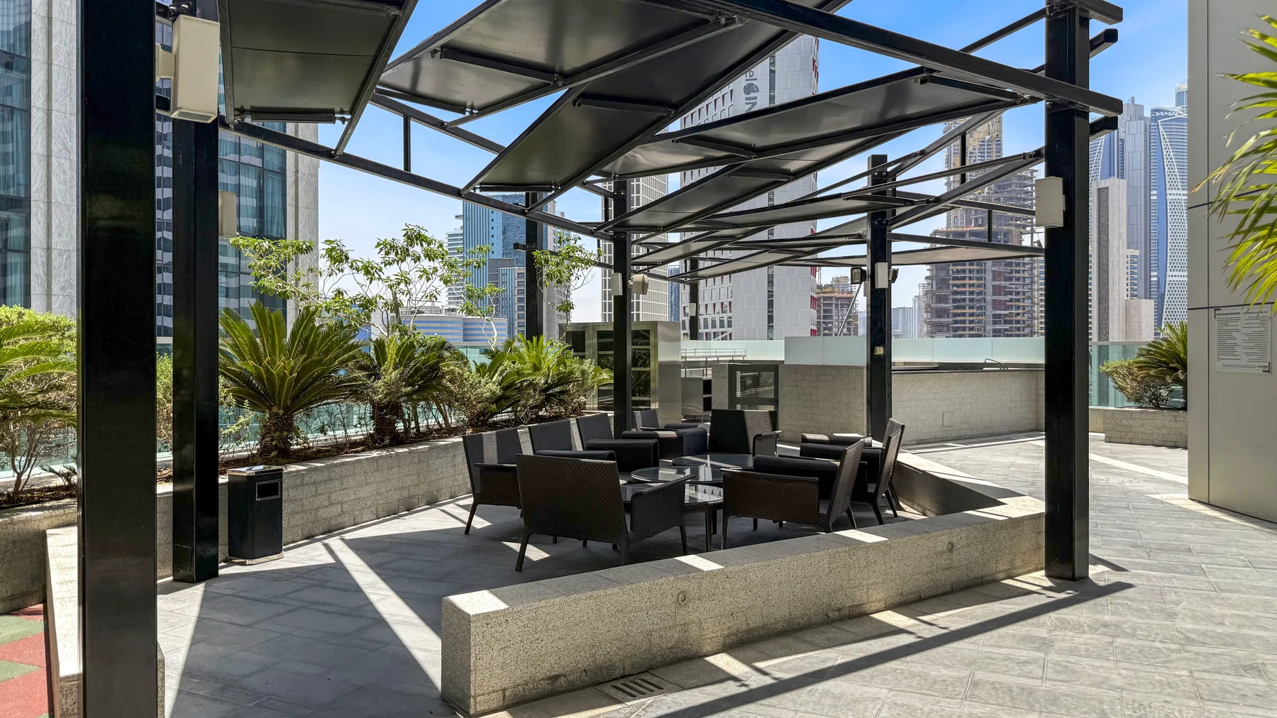 Una terraza en la azotea con asientos al aire libre bajo una cubierta metálica, rodeada de jardineras y edificios.