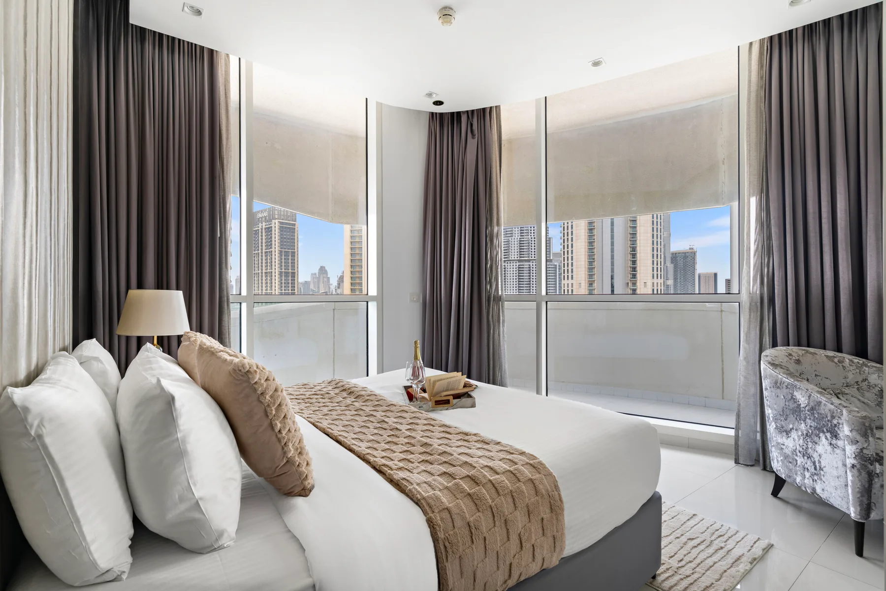 Dormitorio con una cama bien arreglada junto a grandes ventanas con vistas a edificios de la ciudad.
