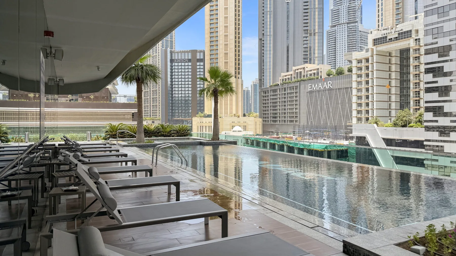 Una piscina al aire libre junto a filas de asientos reclinables, con edificios altos al fondo.
