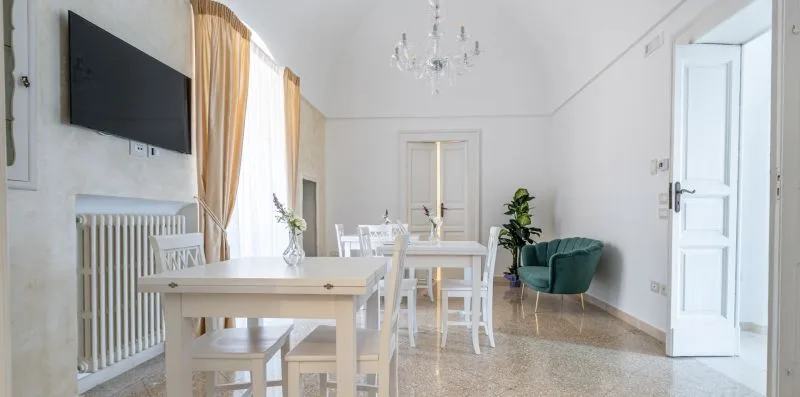 Un comedor luminoso con una mesa, sillas blancas y un televisor montado en la pared.