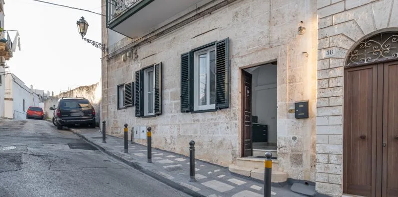 Fachada de piedra con ventanas con persianas y una puerta abierta junto a una calle estrecha.