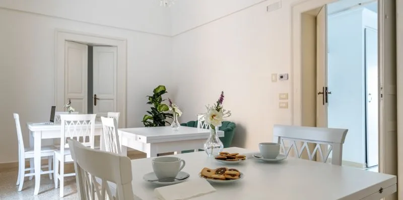 Un comedor luminoso con mesas y sillas blancas, decorado con flores y plantas.