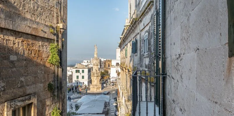 Una calle histórica estrecha se abre a una plaza soleada con edificios de piedra.