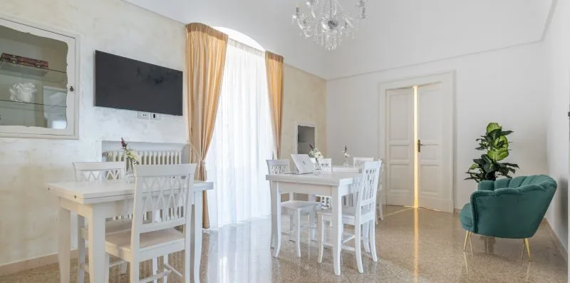Un comedor luminoso con mesas y sillas blancas junto a una ventana con cortinas.