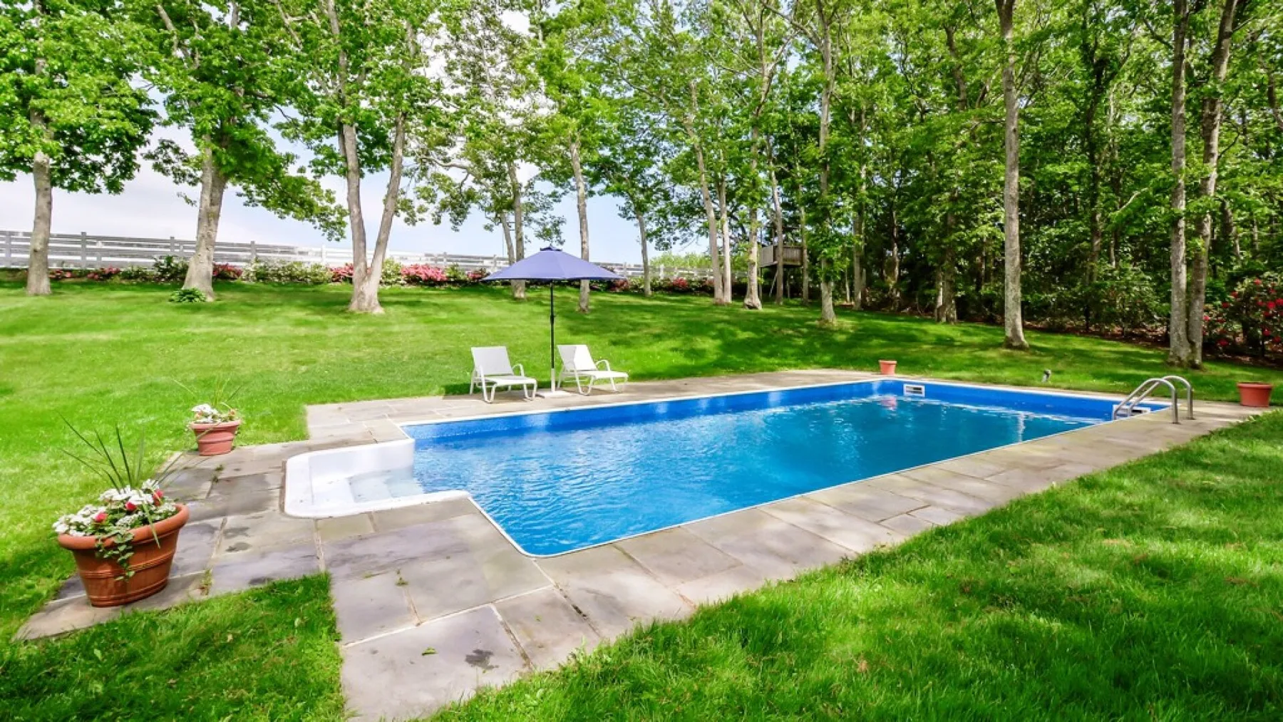 Una piscina al aire libre está en un jardín con césped y árboles frondosos.
