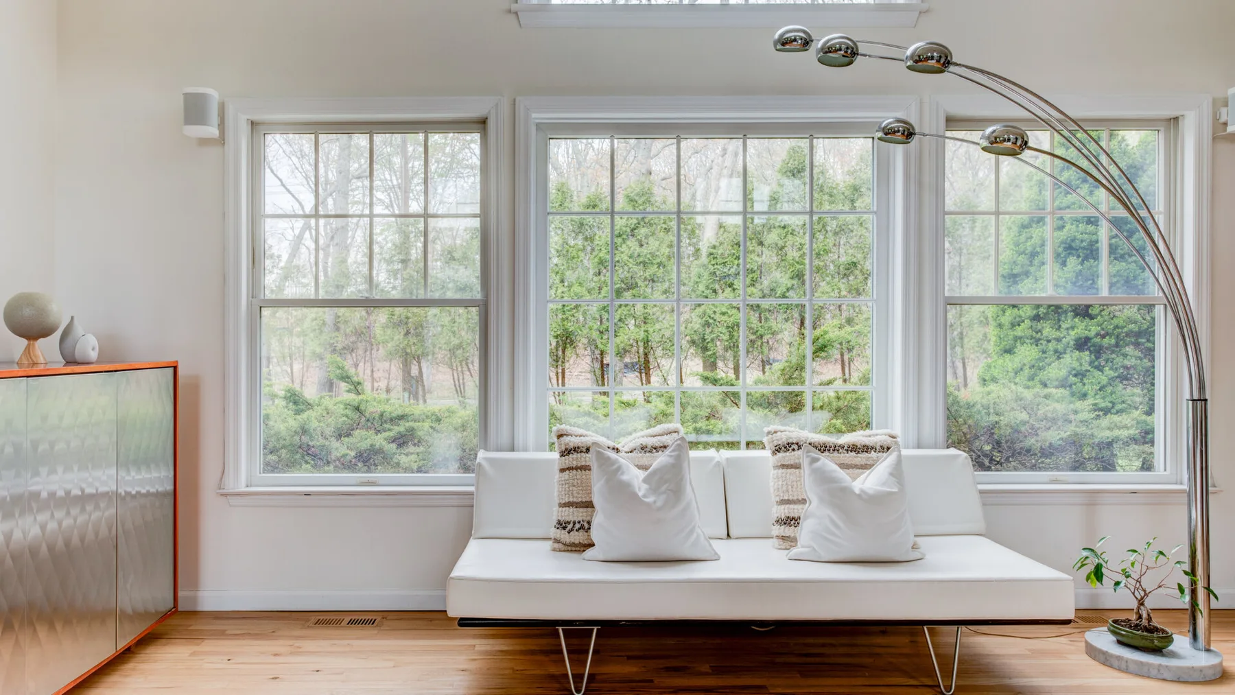 Una sala de estar luminosa con un diván blanco bajo grandes ventanas.
