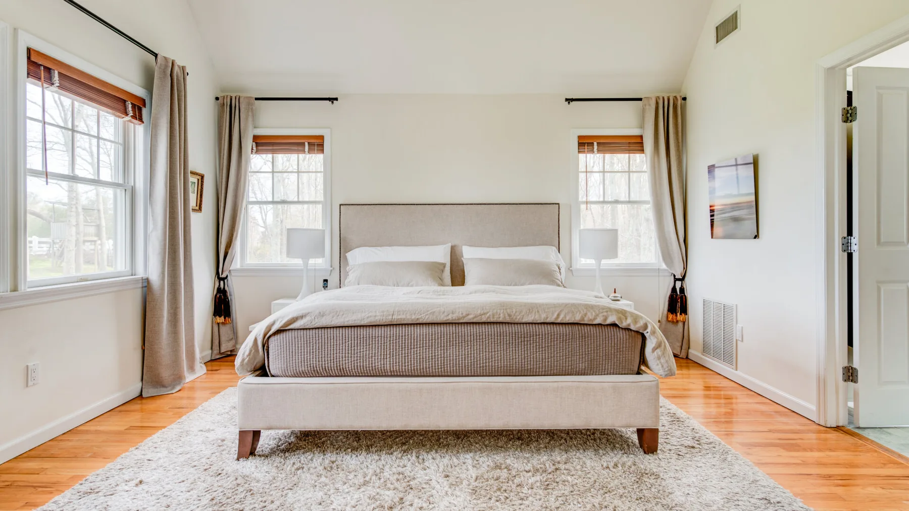Un dormitorio con una cama grande entre dos ventanas y suelo de madera clara.