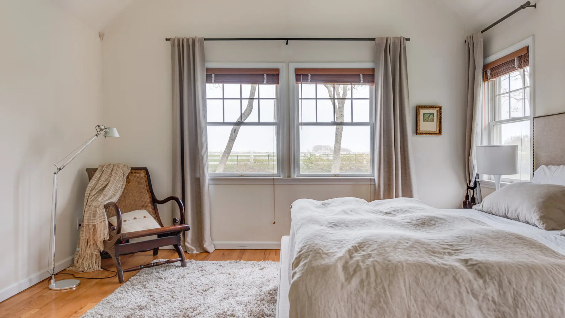 Un dormitorio luminoso con una cama, dos ventanas y una silla de lectura.