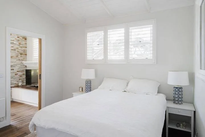 A bright bedroom with a large bed beneath windows fitted with blinds.