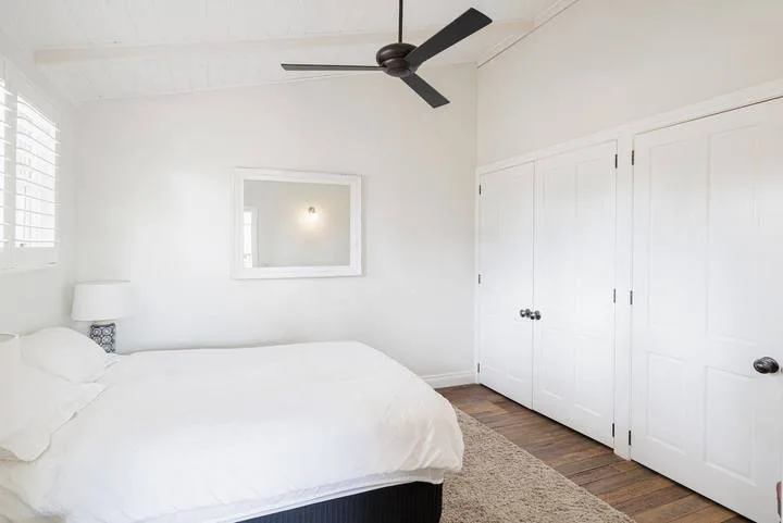 A bright bedroom with a white bed, a wall mirror, and a ceiling fan.