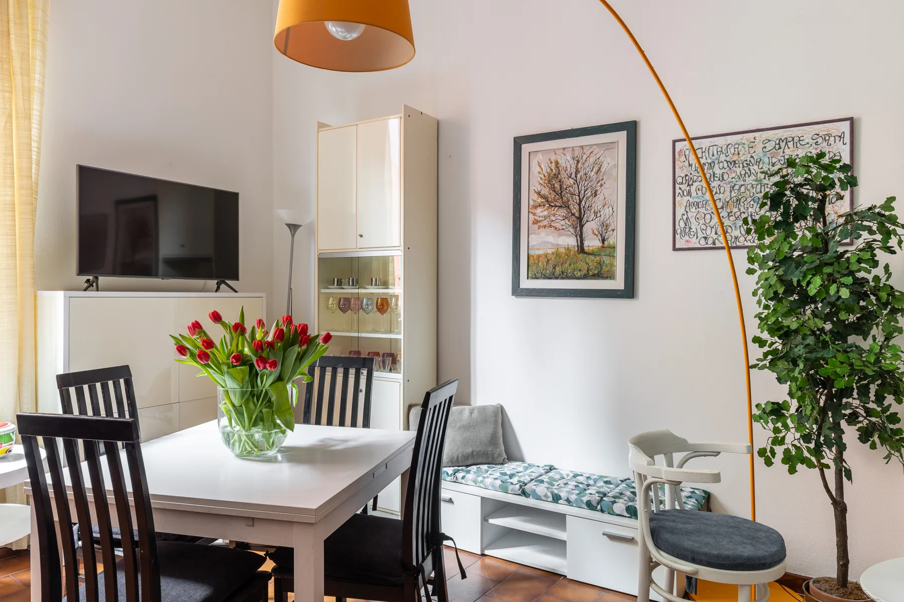 A bright dining and sitting area with a table, framed artwork, and leafy plants.