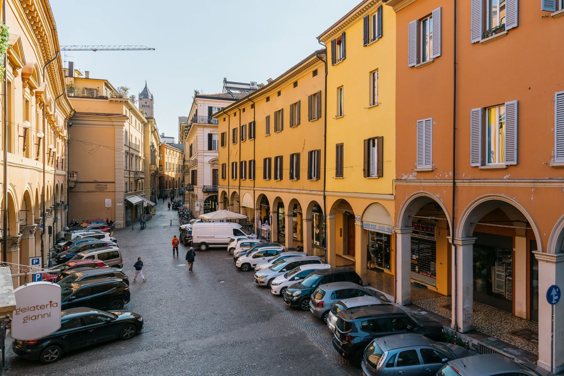 A narrow city street lined with colorful buildings, arched storefronts, and parked cars.