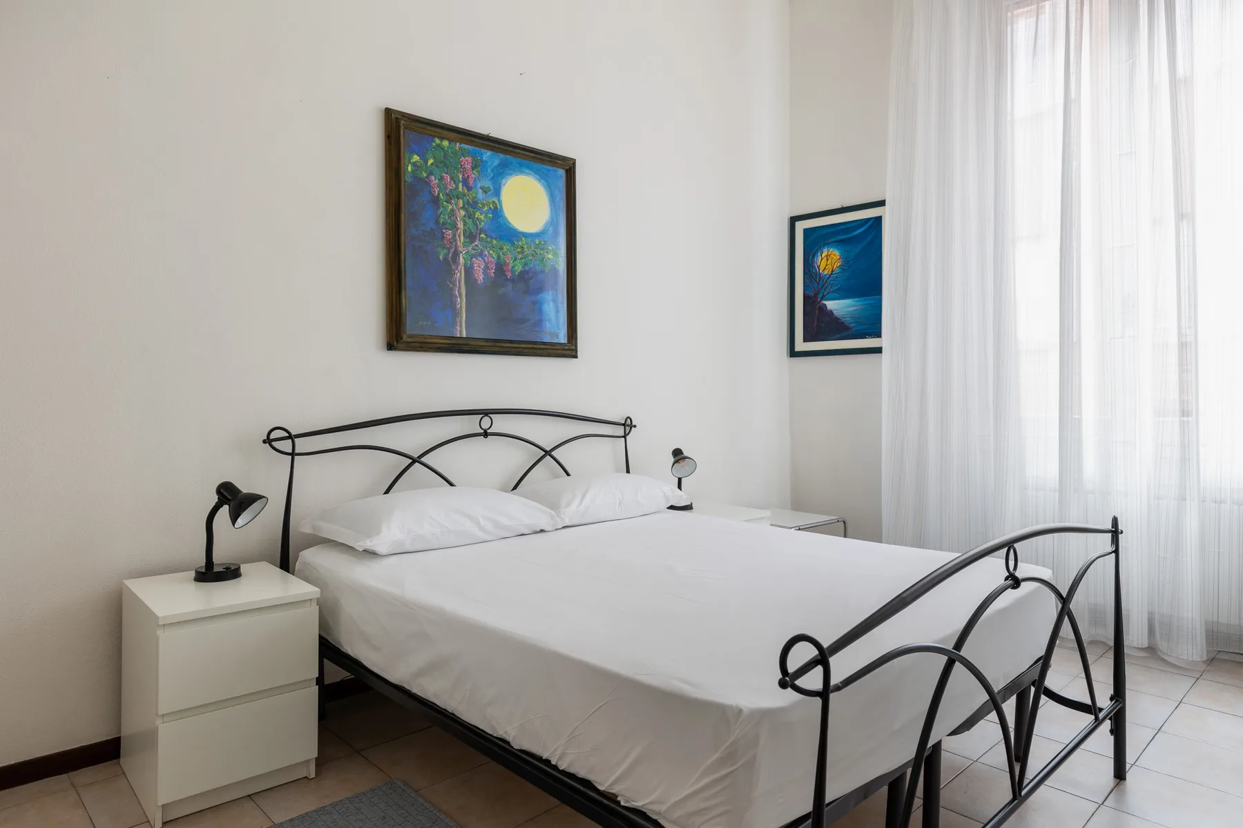 A bright bedroom with a metal bed, white bedding, and large curtained windows.