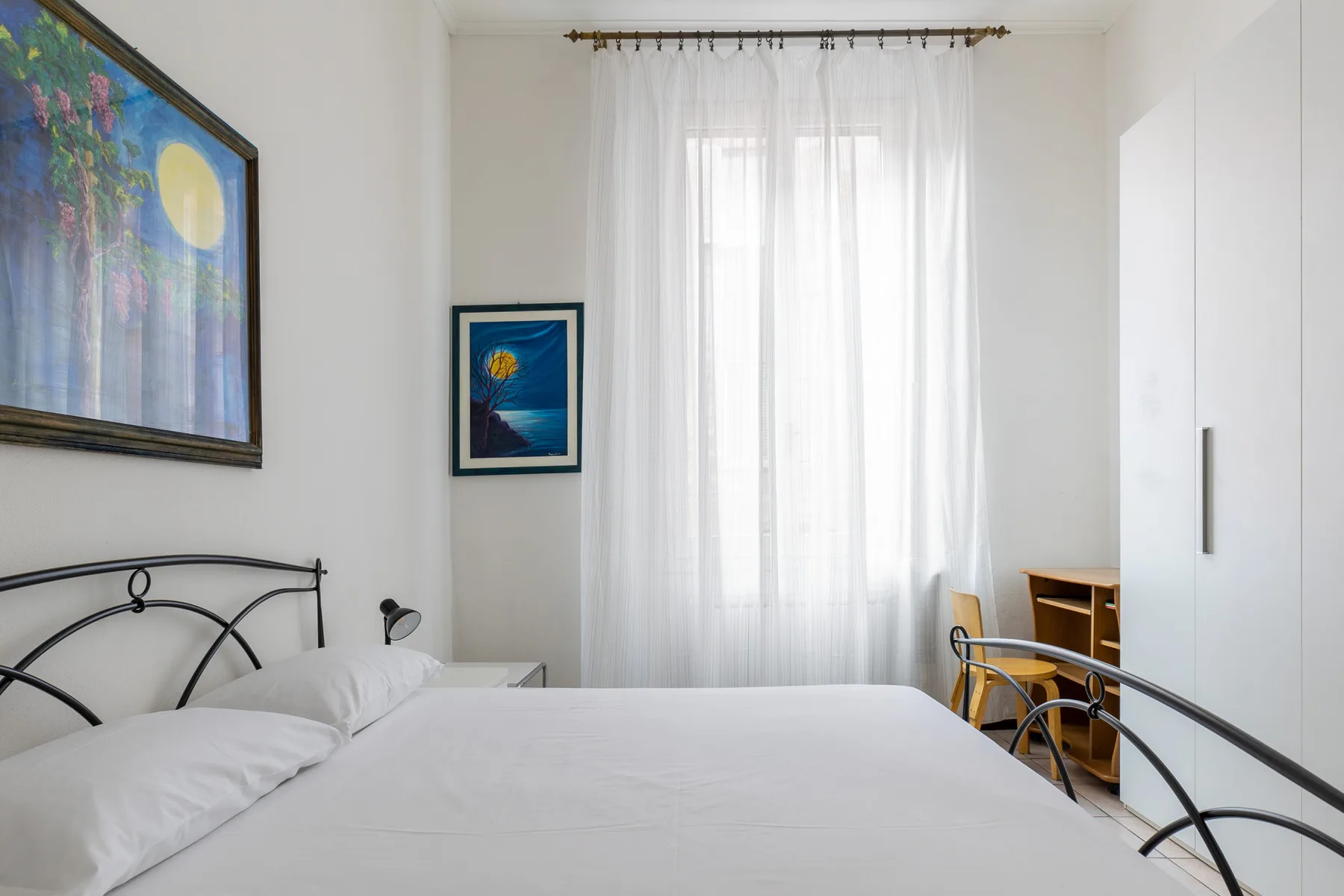 A bright bedroom with a bed, sheer curtains, and framed artwork.