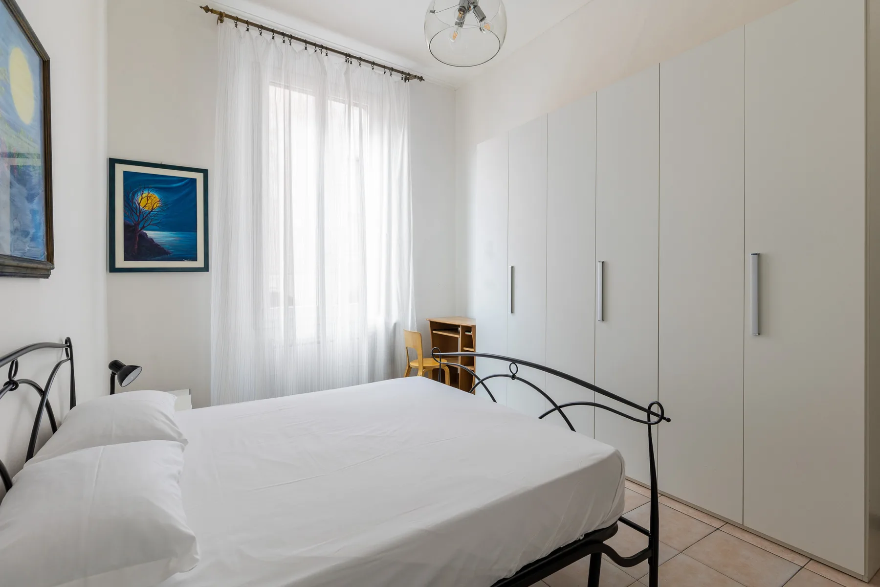 A bright bedroom with a white bed, tall wardrobes, and a curtained window.