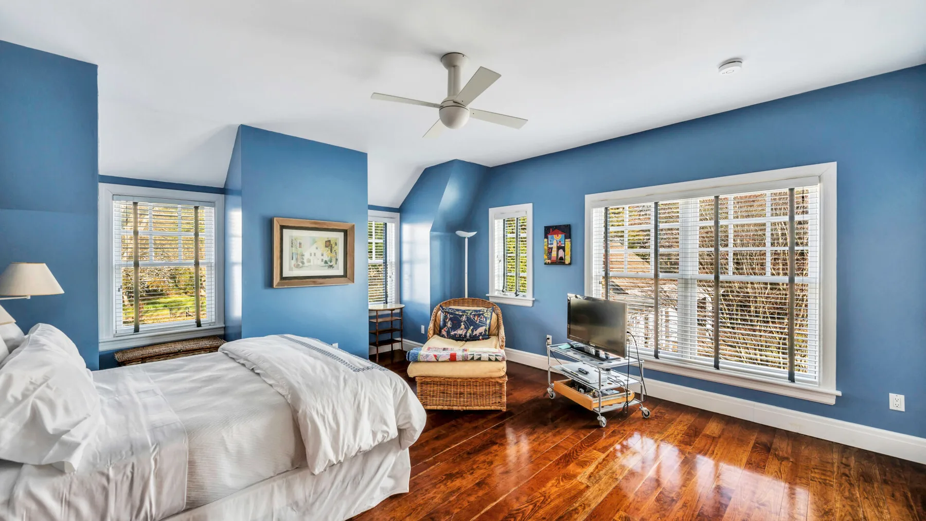 Un dormitorio azul con ropa de cama blanca, un ventilador de techo y ventanas amplias.