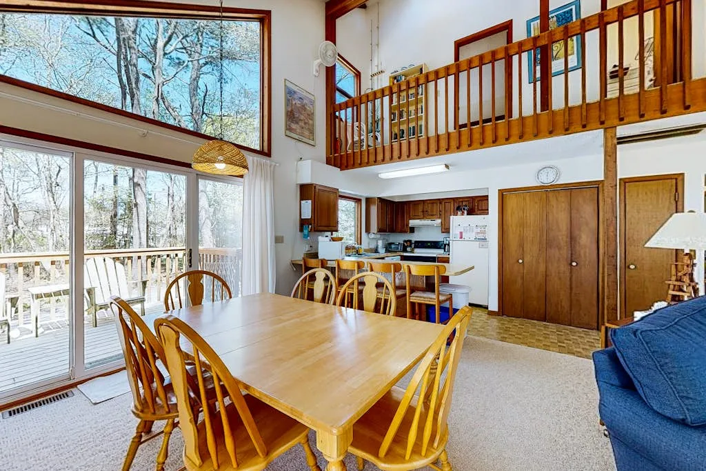 Comedor con una mesa de madera junto a grandes ventanas y una cocina abierta.