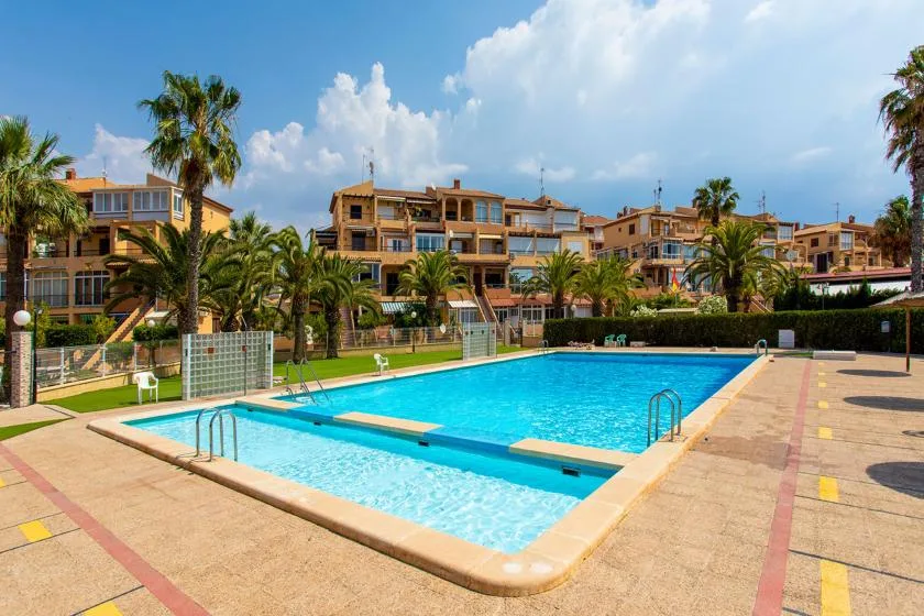 An outdoor swimming pool sits beside a paved deck, with apartment buildings in the background.