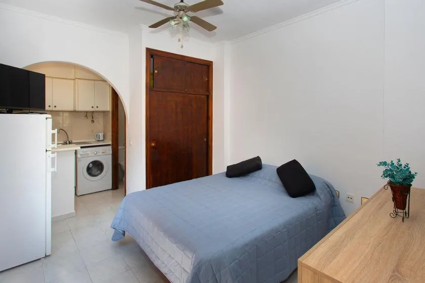 A bedroom with a bed, wooden door, and kitchenette visible through an archway.
