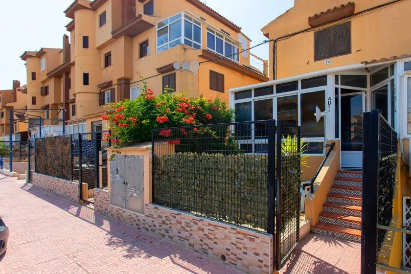 An orange residential building behind a black fence, with a flower-covered shrub by the entrance.