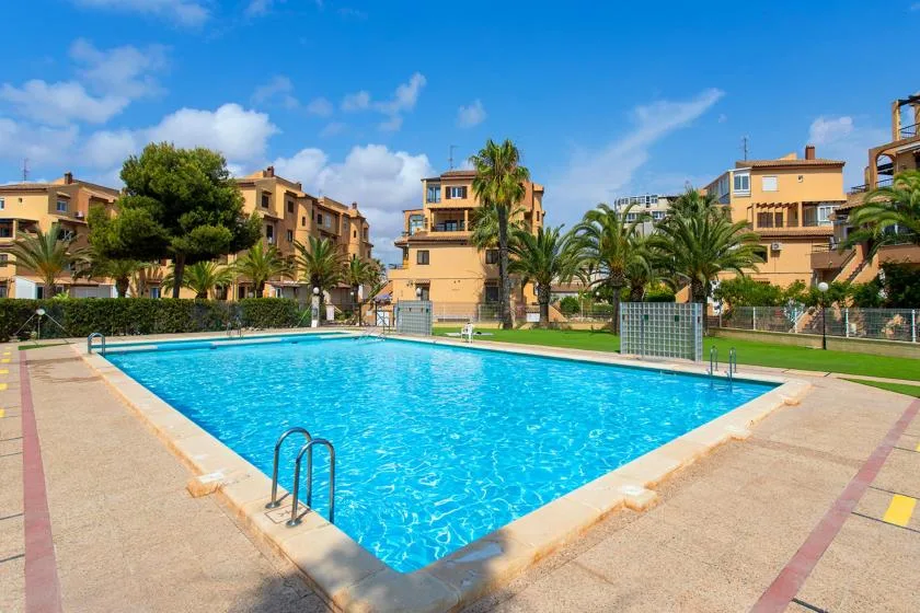 A swimming pool sits among landscaped grounds and apartment buildings under a blue sky.
