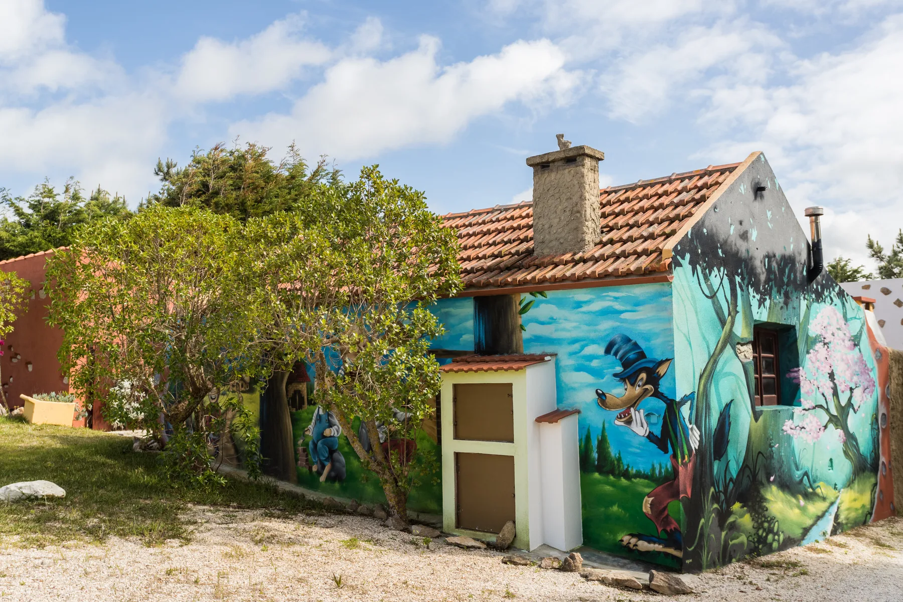 A small house with colorful murals stands beside leafy trees under a partly cloudy sky.