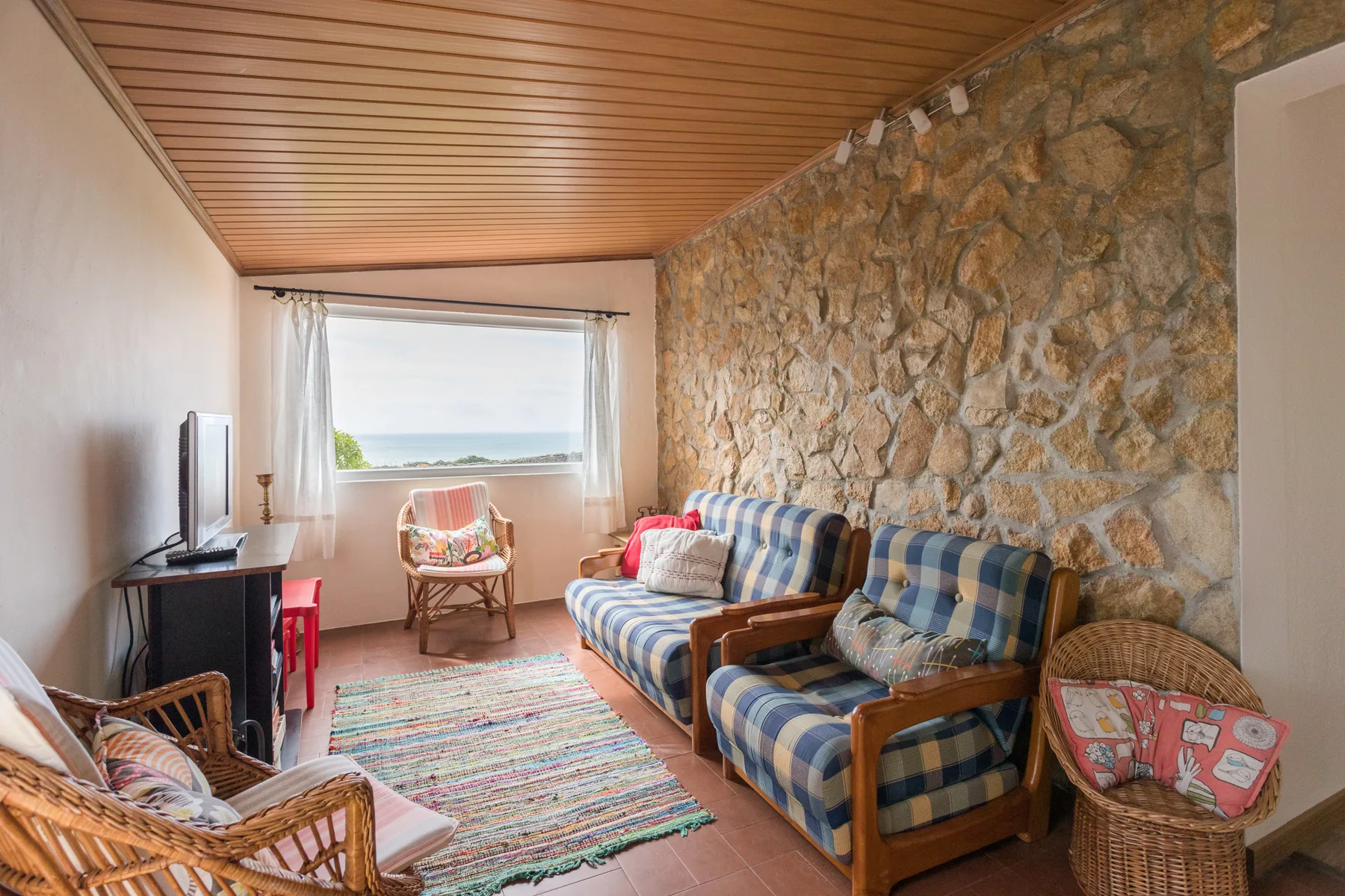 A living room with patterned seating, a television, and a window overlooking the sea.