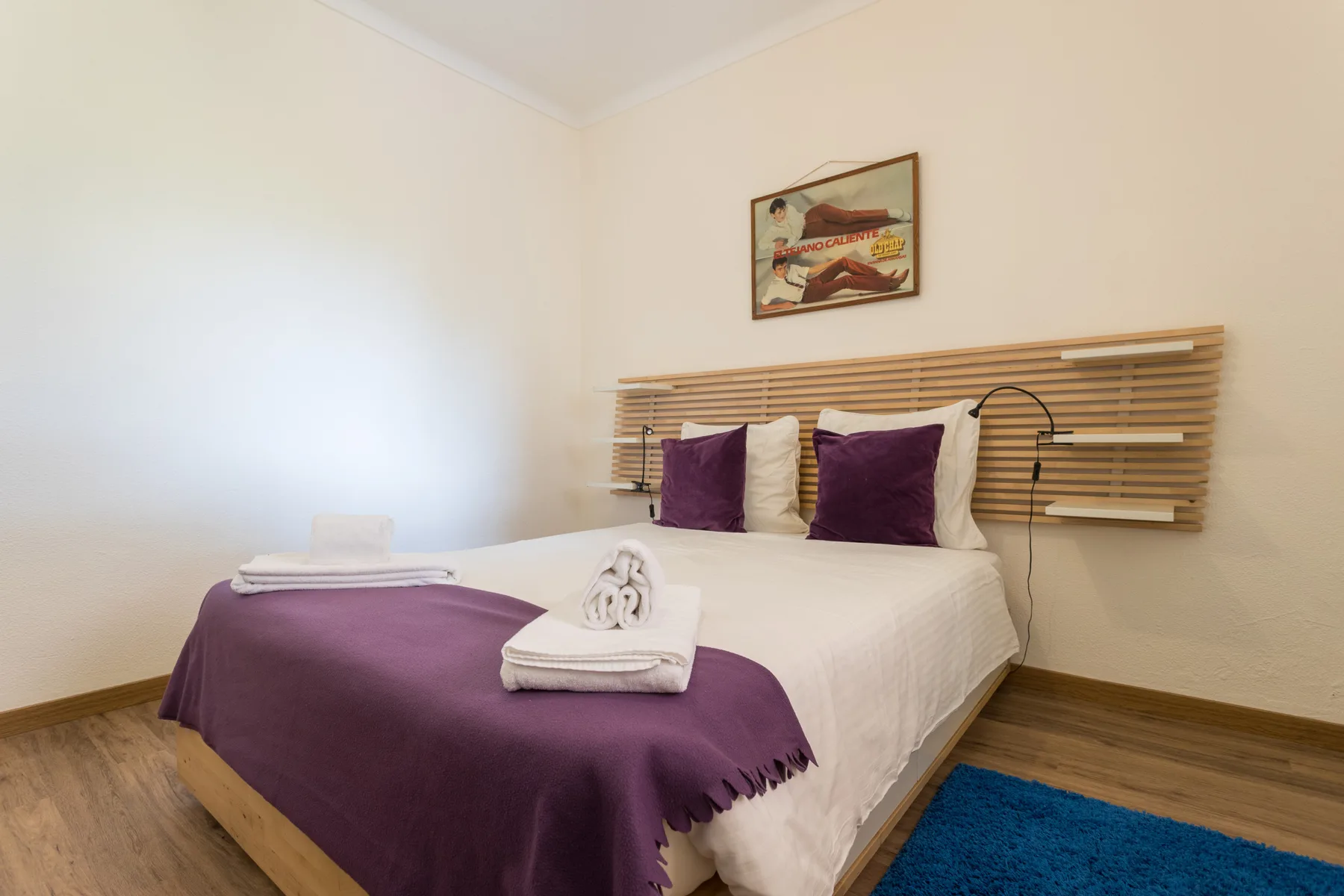 A bedroom with a bed dressed in white linens and purple accents.
