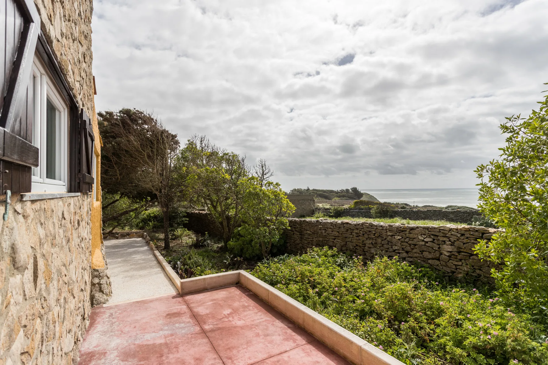 A stone house wall borders a paved terrace overlooking a garden and low stone wall.