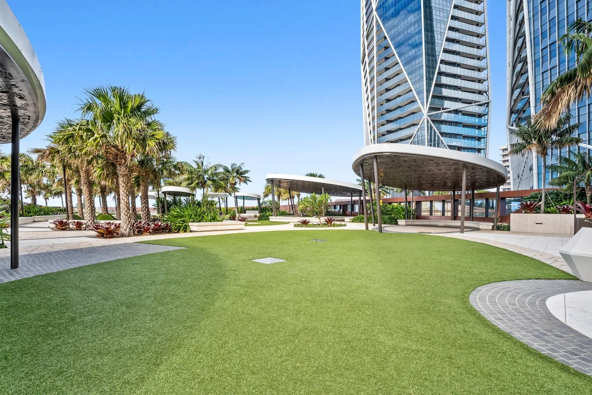 Un patio ajardinado con césped verde, palmeras y edificios modernos de gran altura.