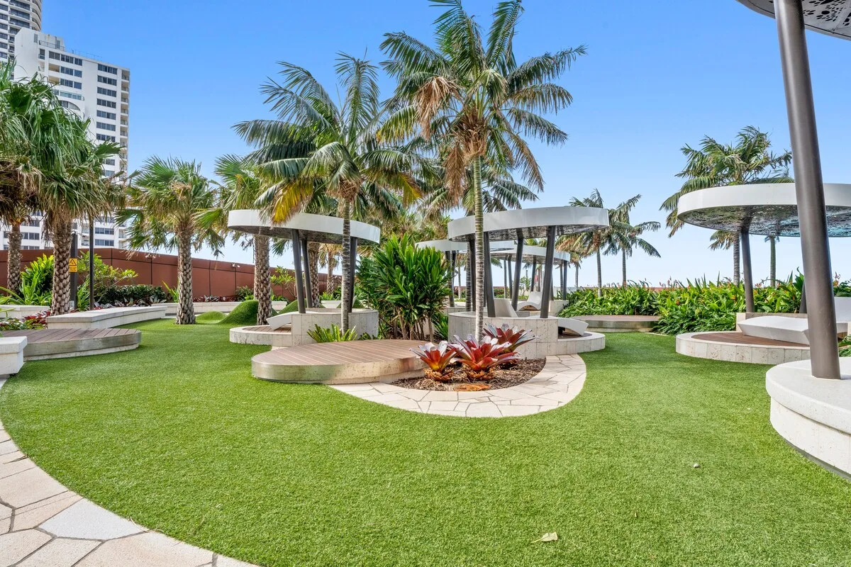 Un jardín exterior ajardinado con palmeras, césped verde y zonas de asientos curvos.