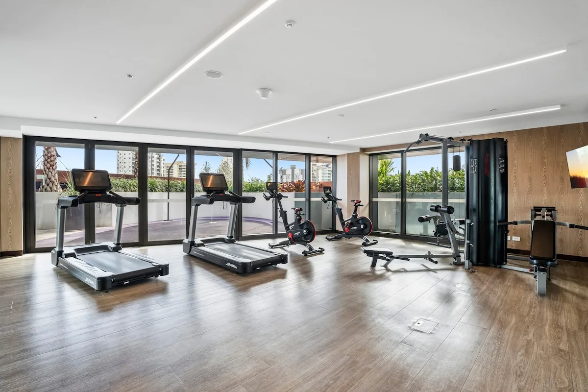 Un gimnasio con cintas de correr y equipo de ejercicio junto a grandes ventanas.