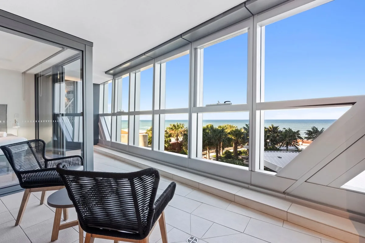 A bright seating area overlooks palm trees and the ocean through large windows.