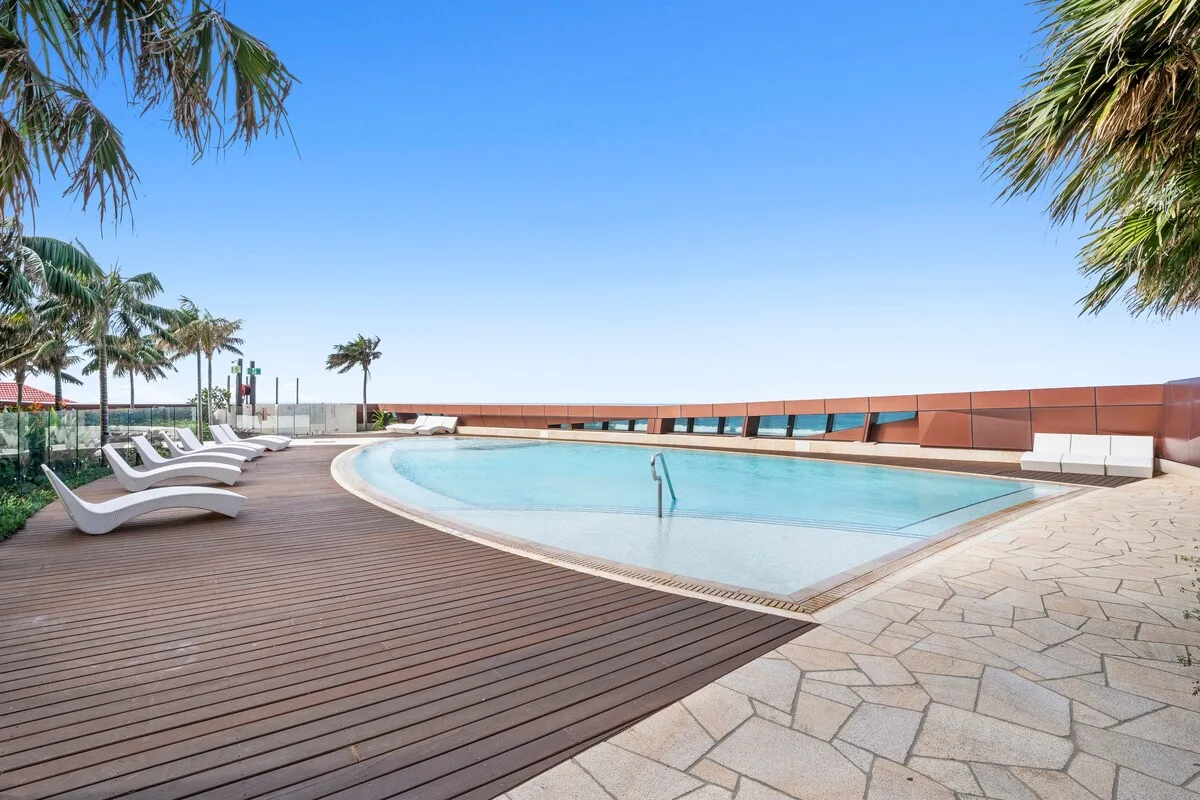 Una piscina curva está junto a una terraza con tumbonas y palmeras.