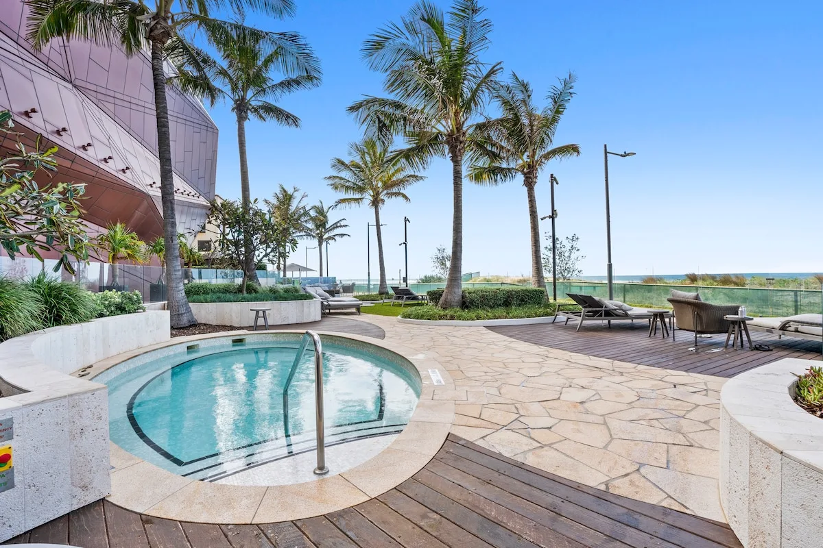 Un patio con piscina, asientos y palmeras junto al mar.