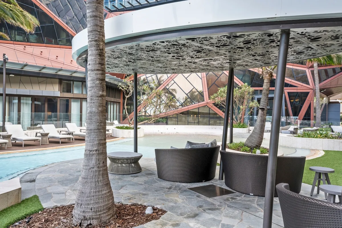 Zona de piscina en un patio con asientos a la sombra y palmeras.