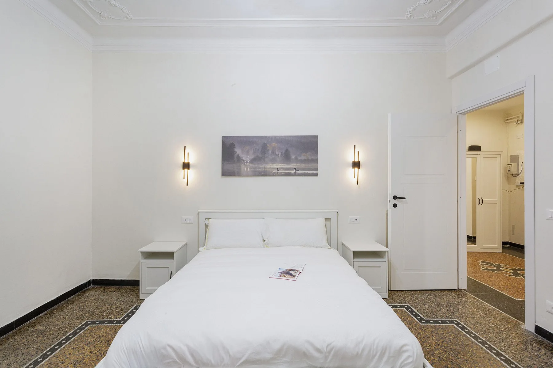Un dormitorio luminoso con una cama tendida y una lámina enmarcada encima.