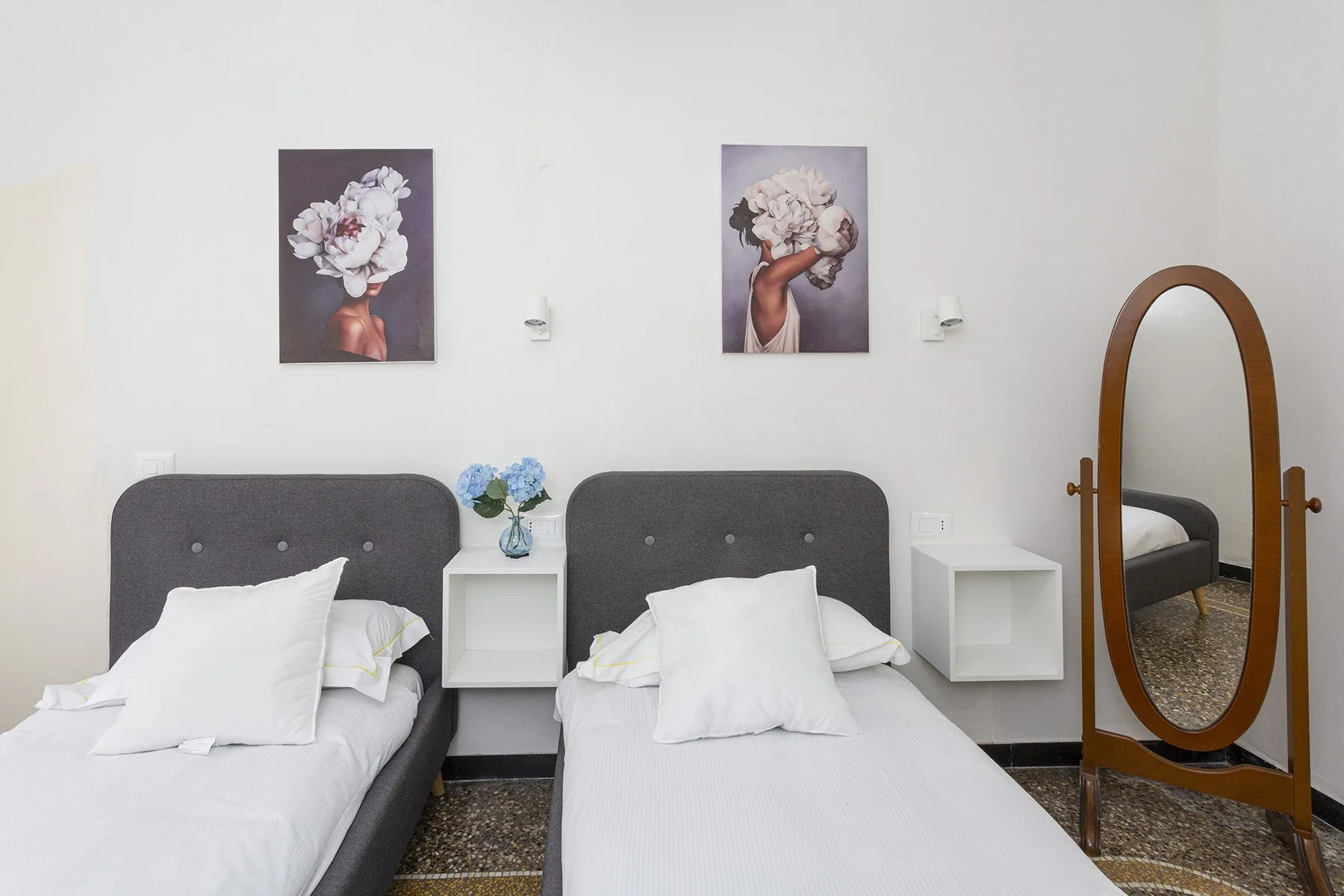 Un dormitorio con dos camas individuales, cuadros florales y un espejo ovalado de pie.