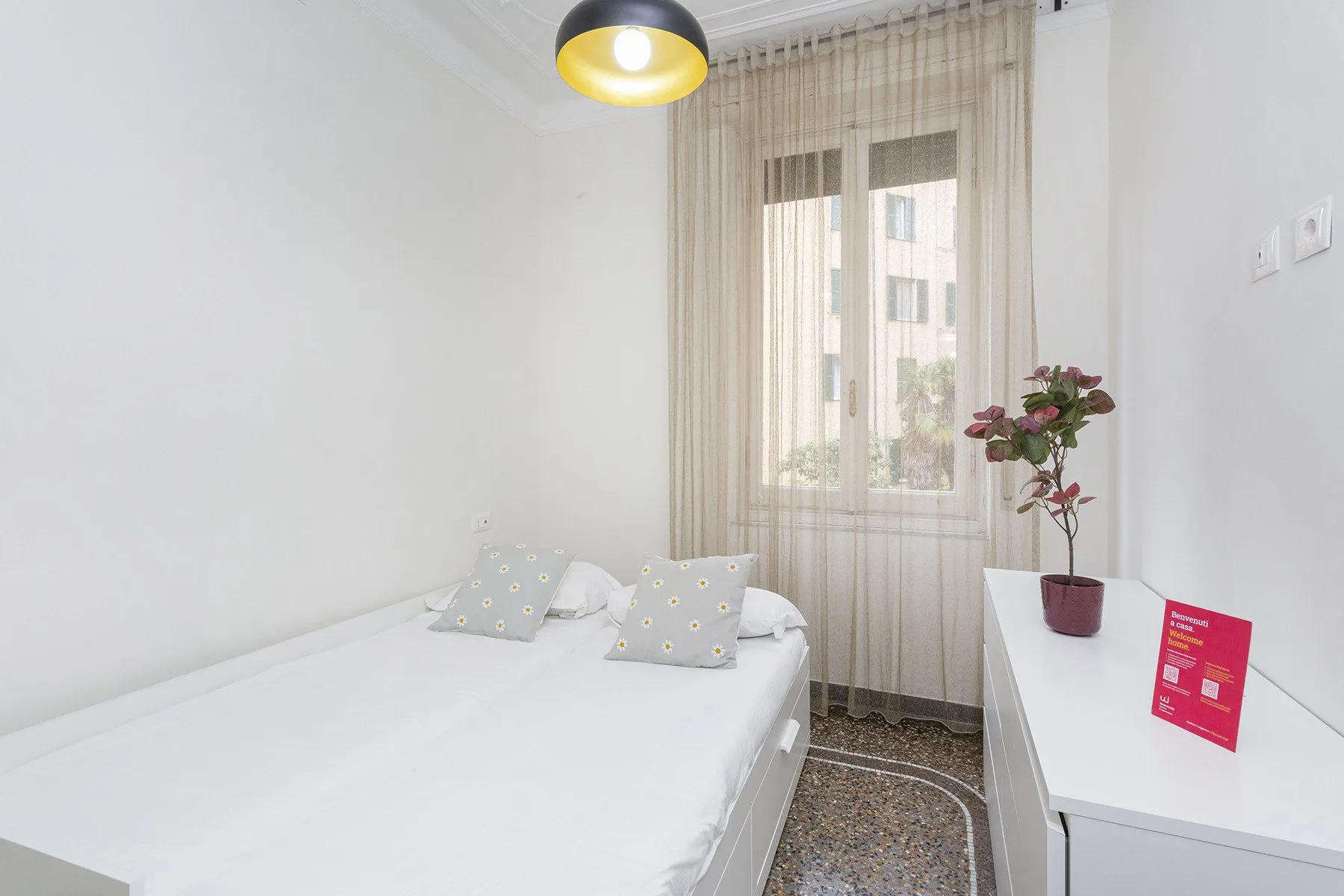 Un dormitorio luminoso con una cama tendida y una ventana con cortinas transparentes.