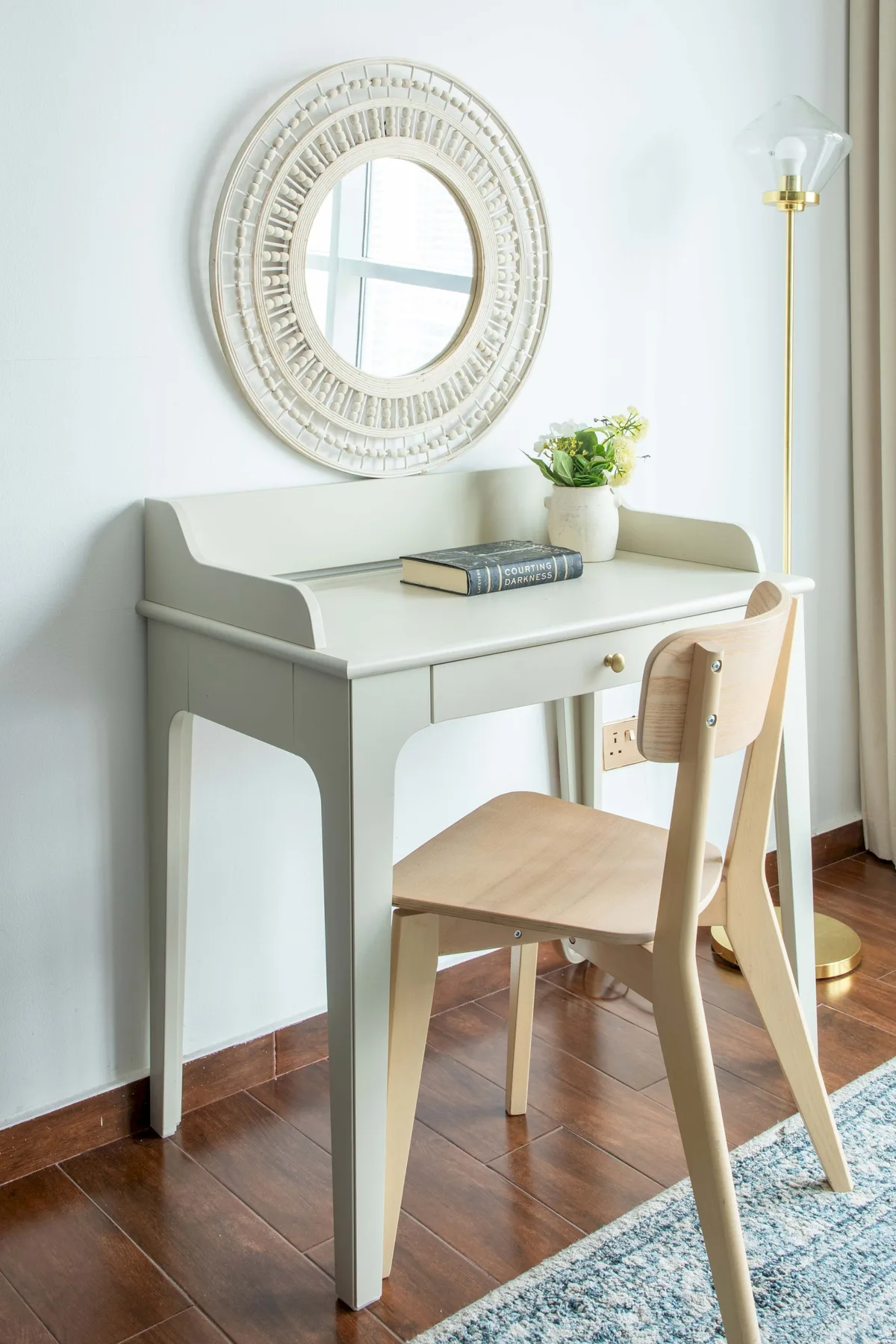 A writing desk and wooden chair sit beneath a round wall mirror.
