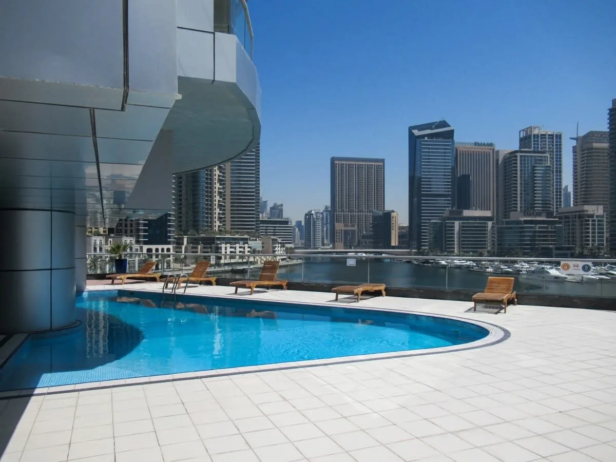 A rooftop pool overlooks a waterfront skyline, with lounge seating beside the water.