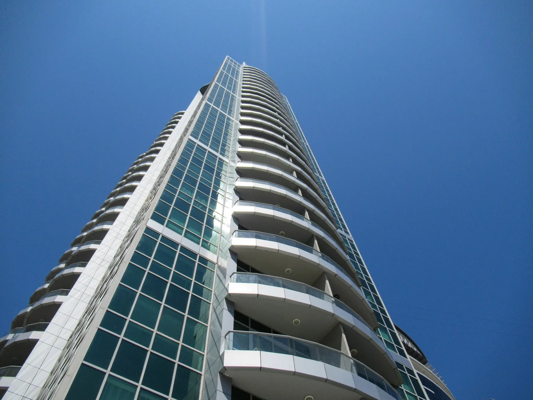 A tall modern tower with glass windows and curved balconies rises against a clear blue sky.