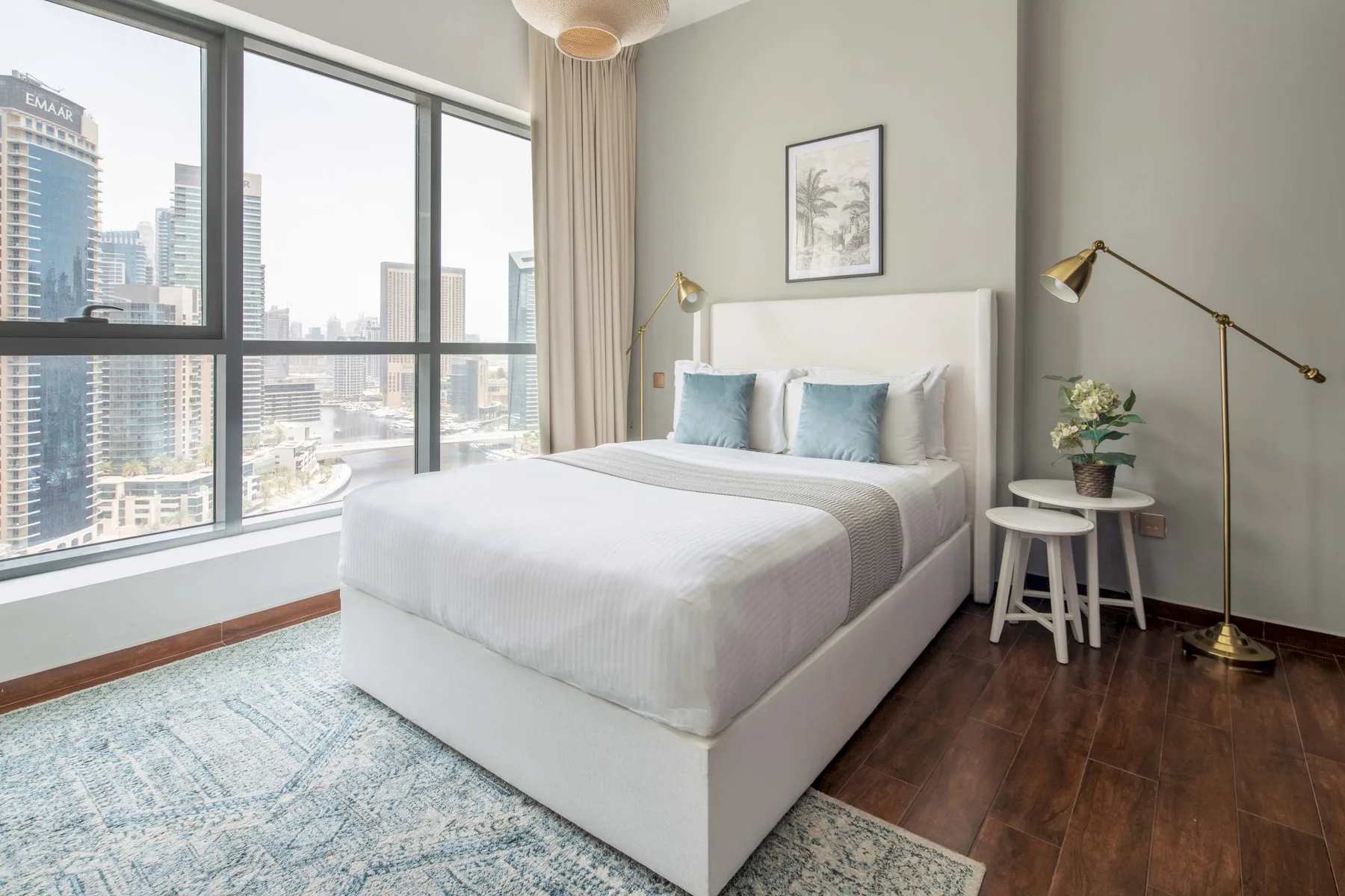 A bright bedroom with a white bed beside large windows overlooking city buildings.