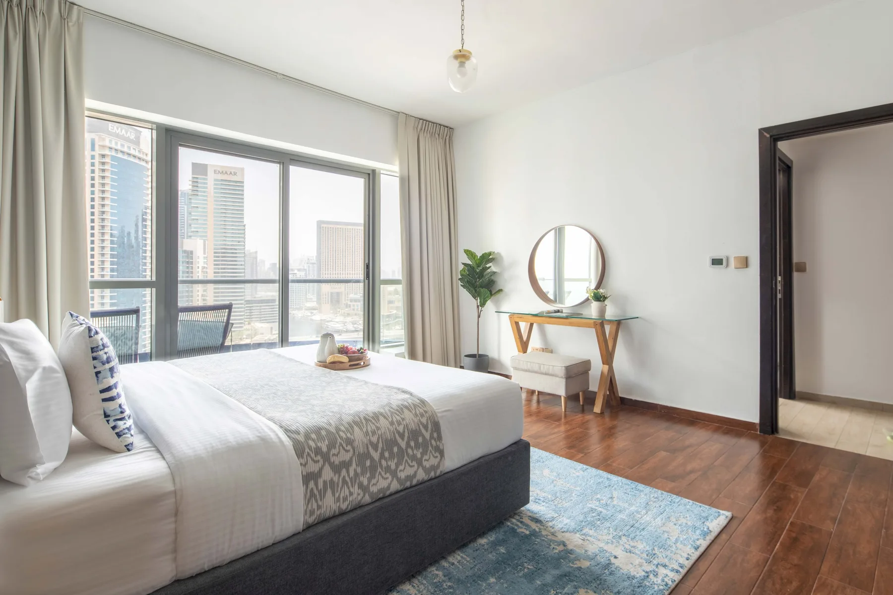 A bright bedroom with a bed beside large windows overlooking nearby buildings.