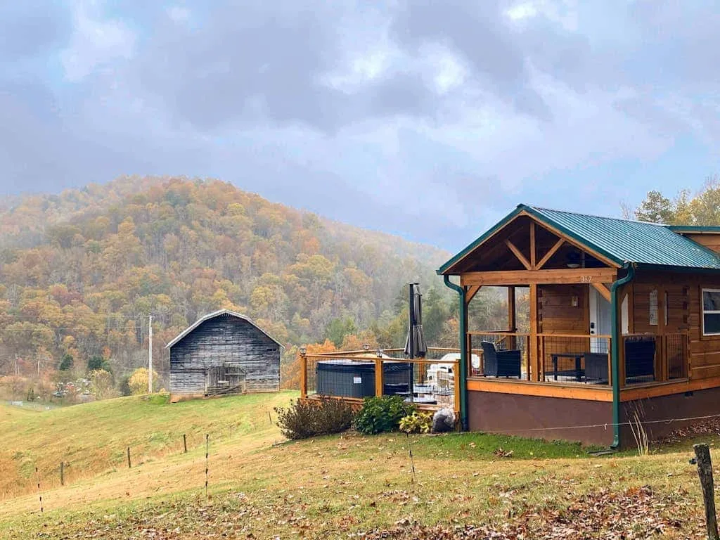 A wooden cabin with a covered porch sits on a grassy hillside beneath wooded hills.