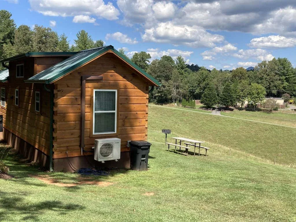 Una pequeña cabaña de madera está junto a una zona abierta de césped bajo un cielo parcialmente nublado.