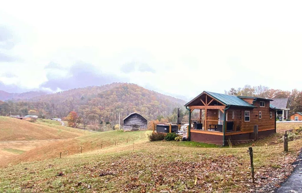 Una cabaña de madera está junto a un campo abierto, con colinas boscosas al fondo.