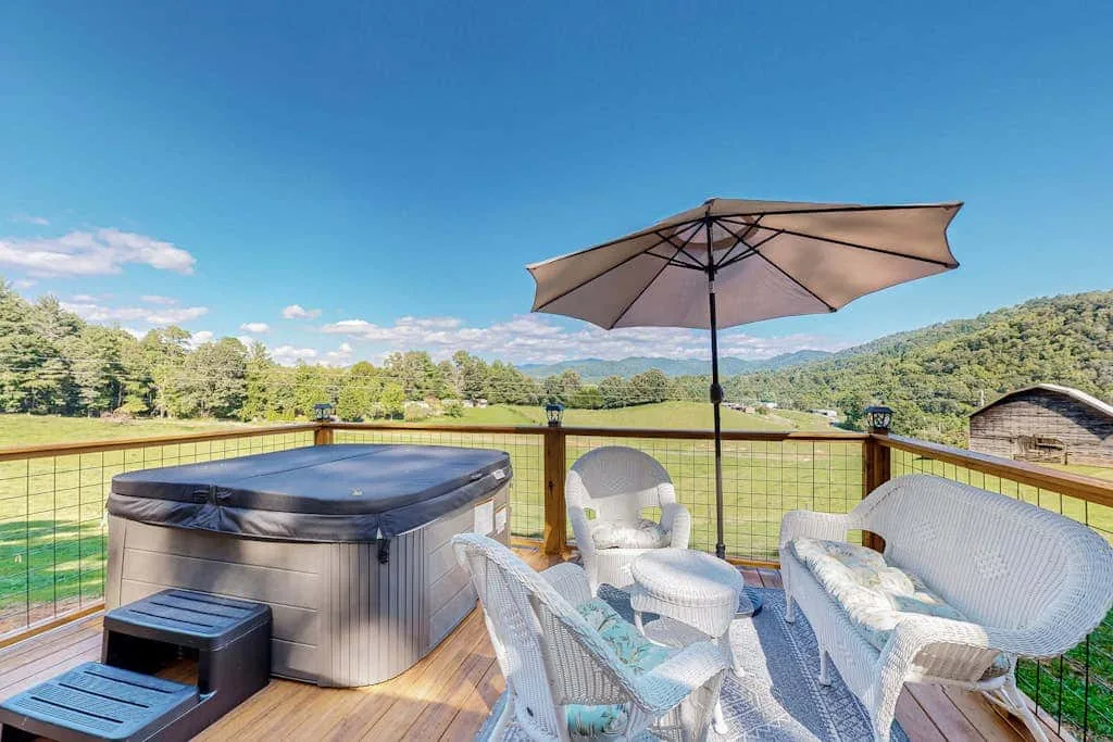 Una terraza exterior tiene una bañera de hidromasaje, asientos blancos y una sombrilla frente a un paisaje verde.