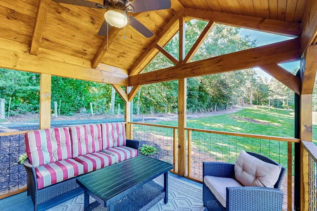 A covered wooden patio has a striped sofa, a table, and seating overlooking greenery.